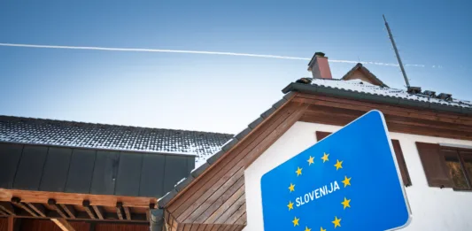 A blue roadside panel with the 12 stars of the European Union and the word 'Slovenija' marks the entrance to Slovenia on the route from Austria. The sign stands near buildings of a former checkpoint in a mountain setting, illustrating the Schengen system where routine border controls have been removed and movement is simplified for travelers. This clear, close composition is useful for stories on EU membership, mobility, tourism flows, cross-border commuting, transport policy, and the visual language of European road signage at internal borders.