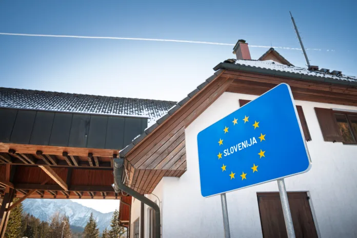a-blue-roadside-panel-with-the-12-stars-of-the-european-union-and-the-word-slovenija-marks-the-entrance-to-slovenia-on-the-route-from-austria-the-sign-stands-near-buildings-of-a-former-checkpoint-in-a-stockpack-istock A blue roadside panel with the 12 stars of the European Union and the word 'Slovenija' marks the entrance to Slovenia on the route from Austria. The sign stands near buildings of a former checkpoint in a mountain setting, illustrating the Schengen system where routine border controls have been removed and movement is simplified for travelers. This clear, close composition is useful for stories on EU membership, mobility, tourism flows, cross-border commuting, transport policy, and the visual language of European road signage at internal borders.