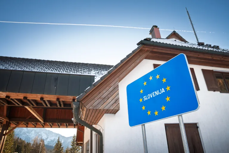 A blue roadside panel with the 12 stars of the European Union and the word 'Slovenija' marks the entrance to Slovenia on the route from Austria. The sign stands near buildings of a former checkpoint in a mountain setting, illustrating the Schengen system where routine border controls have been removed and movement is simplified for travelers. This clear, close composition is useful for stories on EU membership, mobility, tourism flows, cross-border commuting, transport policy, and the visual language of European road signage at internal borders.