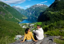 5 destinacij v Severni Evropi, ki vas bodo ohladile in osupnile A couple sits on a rocky outcrop, admiring the stunning fjord landscape of Geiranger Norway.