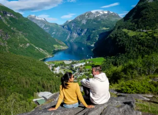 5 destinacij v Severni Evropi, ki vas bodo ohladile in osupnile A couple sits on a rocky outcrop, admiring the stunning fjord landscape of Geiranger Norway.