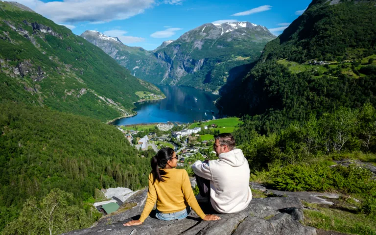 A couple sits on a rocky outcrop, admiring the stunning fjord landscape of Geiranger Norway.
