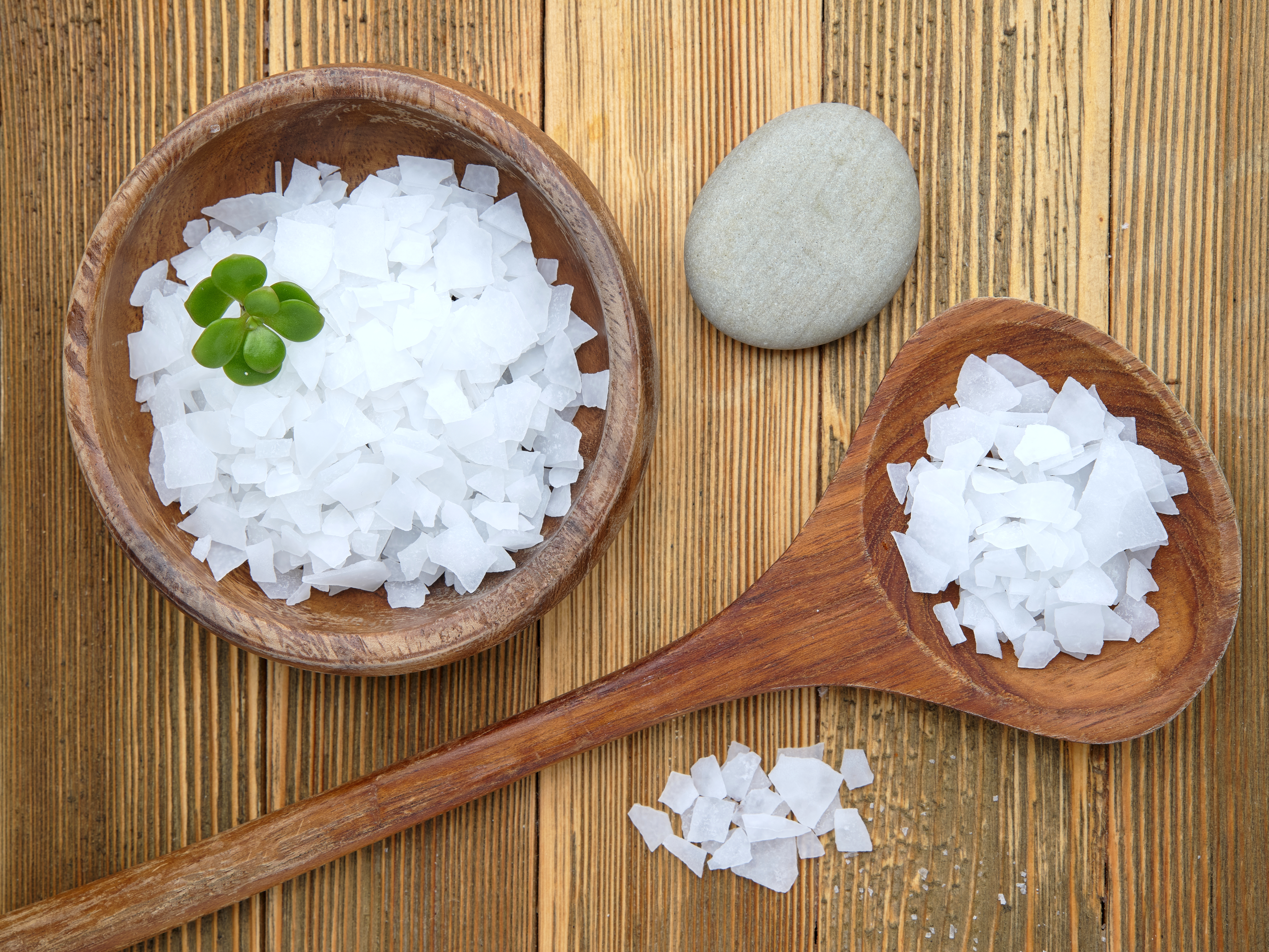 A flat lay photo of magnesium chloride flakes in a wooden bowl and spoon on a wooden table.