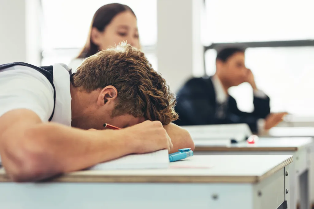 Bored male high school student sleeping during the lecture in the classroom. Schoolboy sleeping on desk in classroom with classmates in background.