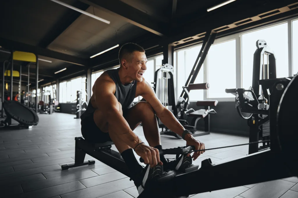 Cheerful attractive man smiling while using rowing machine in contemporary gym. Fit young man working out on rowing equipment at fitness center. Weight loss and cardio exercise concept