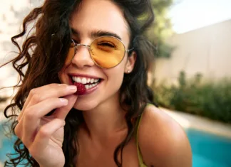 Nosite sončna očala? Strokovnjaki pravijo, da bi jih morali veliko pogosteje Close-up of a beautiful young woman in sunglasses about to eat a raspberry. Outdoor summer scene with pool in background. Healthy lifestyle and freshness.