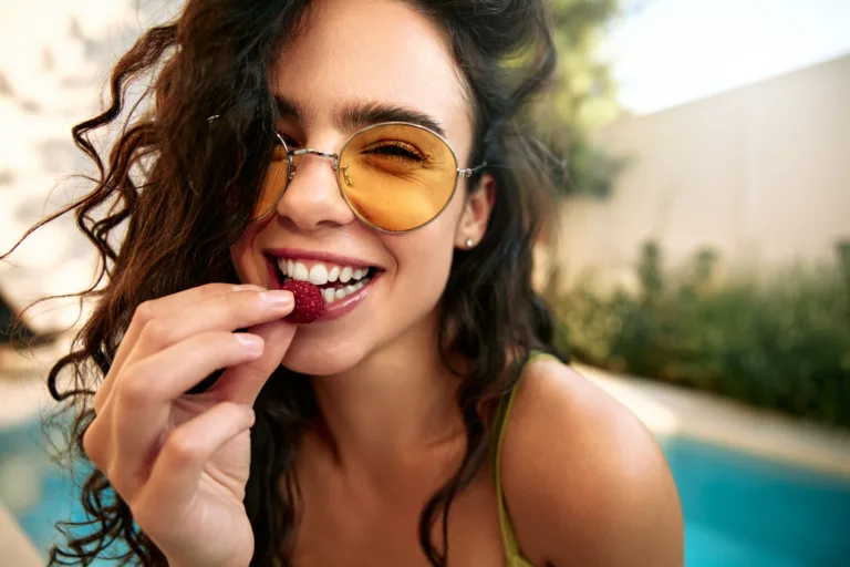 Close-up of a beautiful young woman in sunglasses about to eat a raspberry. Outdoor summer scene with pool in background. Healthy lifestyle and freshness.