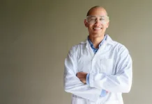 H-indeks: kaj pomeni v akademskem svetu in kolikšen h-indeks označuje uspešnega raziskovalca? Confident male scientist in white lab coat and protective glasses posing against plain wall. Studio science concept.