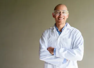 H-indeks: kaj pomeni v akademskem svetu in kolikšen h-indeks označuje uspešnega raziskovalca? Confident male scientist in white lab coat and protective glasses posing against plain wall. Studio science concept.