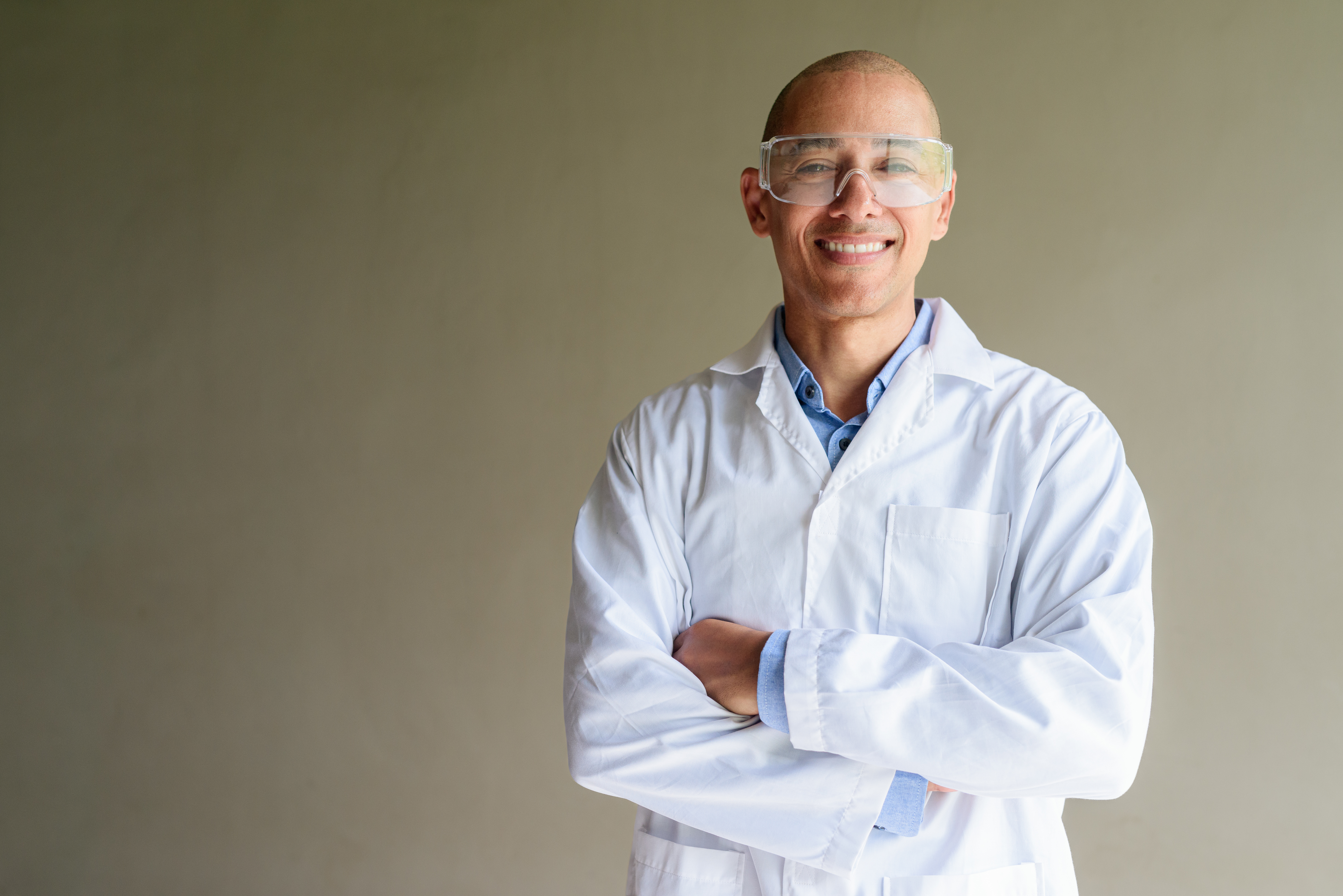 H-indeks: kaj pomeni v akademskem svetu in kolikšen h-indeks označuje uspešnega raziskovalca? Confident male scientist in white lab coat and protective glasses posing against plain wall. Studio science concept.