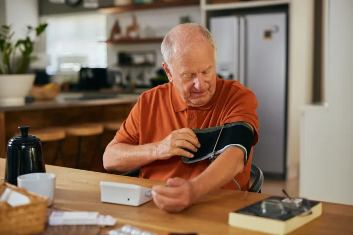 Elderly man checking his blood pressure at home with a digital device. Senior man monitoring health using a blood pressure machine. Serious older man sitting and measuring blood pressure alone.