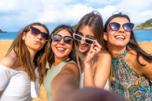 Four young women laughing and posing for a sunny beach selfie, wearing sunglasses and casual summer clothes, enjoying friendship, relaxation and carefree vacation vibes