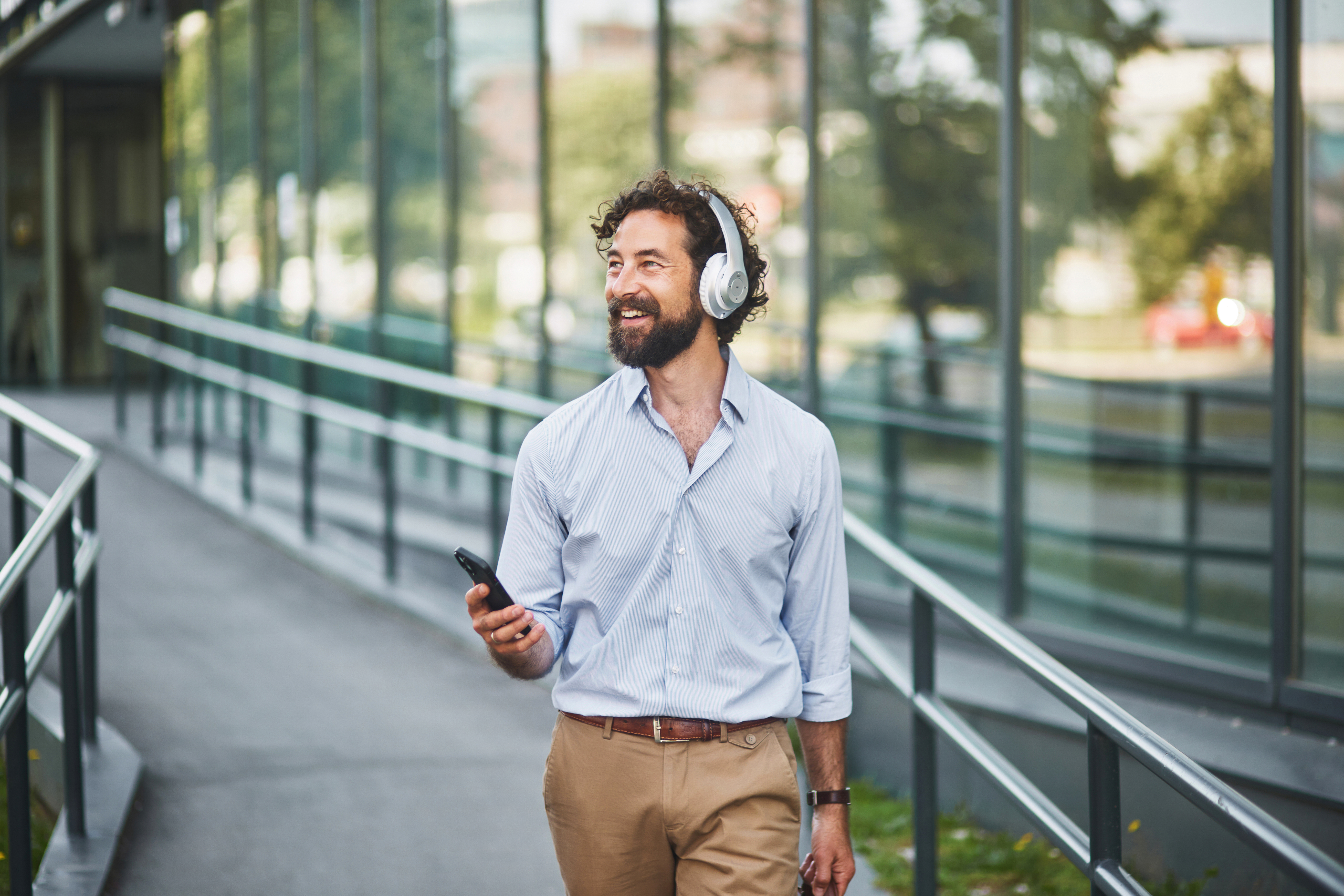 Happy businessman is walking outside the office, holding his smartphone and listening to music