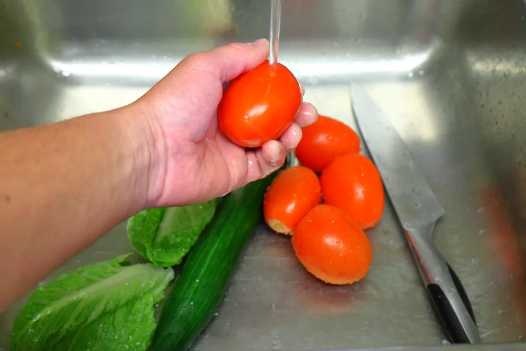 Washing a tomato under running water in a sink with cucumber, lettuce, and a knife nearby.