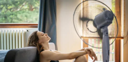 Young woman suffering from heat sitting at home on the floor in front of a fan
