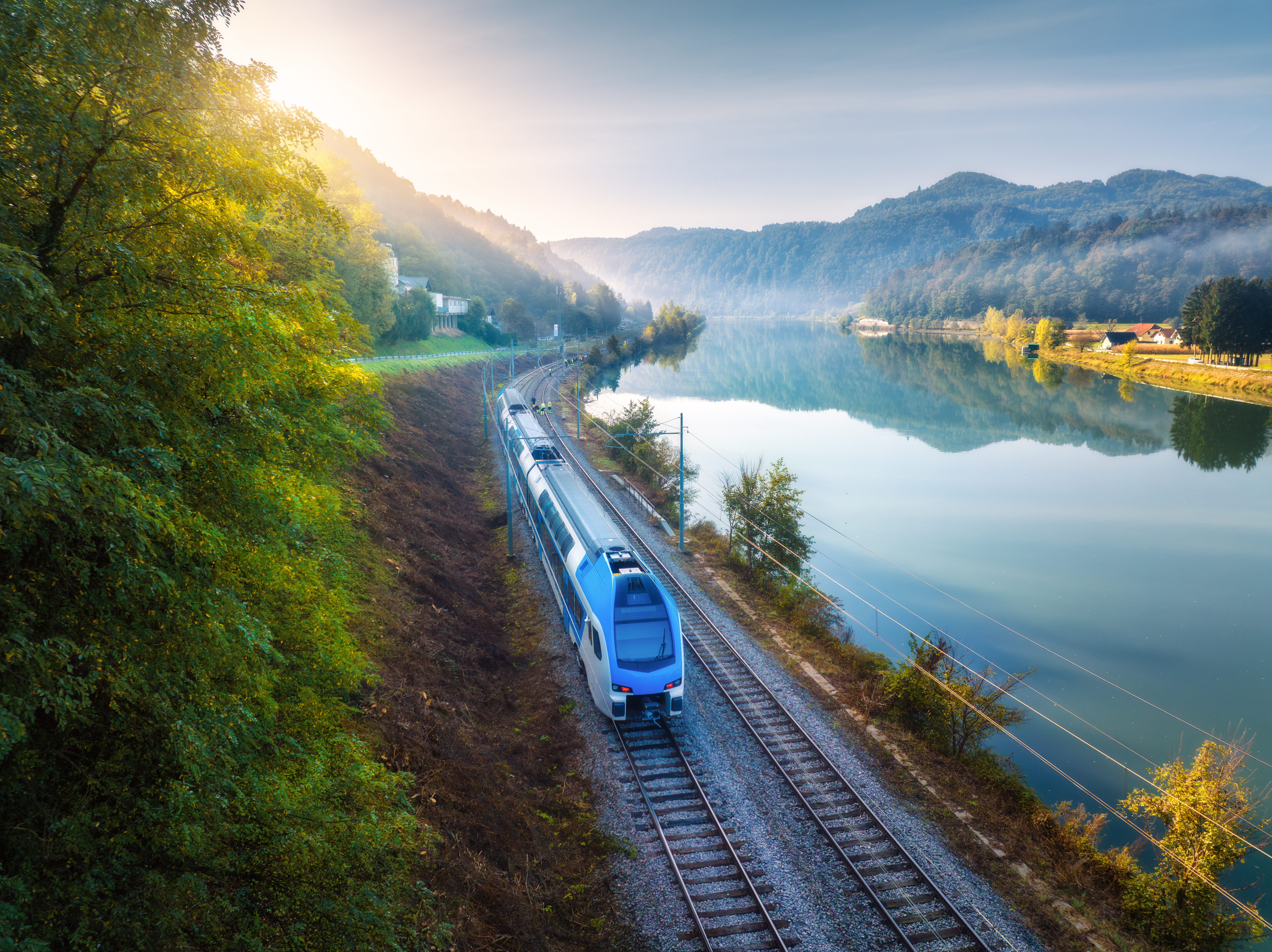 Aerial view of blue modern high speed train moving near river in alpine mountains in fog at sunrise in autumn. Top view of train, railroad, lake, reflection, trees in fall. Railway station in Slovenia