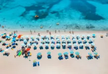 Na italijanskih plažah veljajo nova pravila: če jih ne poznate, vas lahko drago stanejo Aerial view of colorful umbrellas on sandy beach, swimming people in blue sea at summer sunny day. Sardinia, Italy. Tropical seascape. Travel and vacation. Top drone view of ocean with azure water