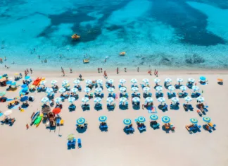 Na italijanskih plažah veljajo nova pravila: če jih ne poznate, vas lahko drago stanejo Aerial view of colorful umbrellas on sandy beach, swimming people in blue sea at summer sunny day. Sardinia, Italy. Tropical seascape. Travel and vacation. Top drone view of ocean with azure water
