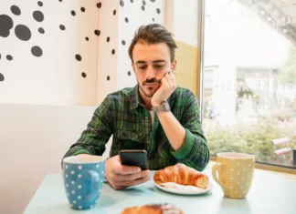 Tinder je uradno iz mode: Zakaj se mladi vračajo k spoznavanju v živo? Bearded hipster man sitting at table in a cafe having breakfast. Guy using smartphone.