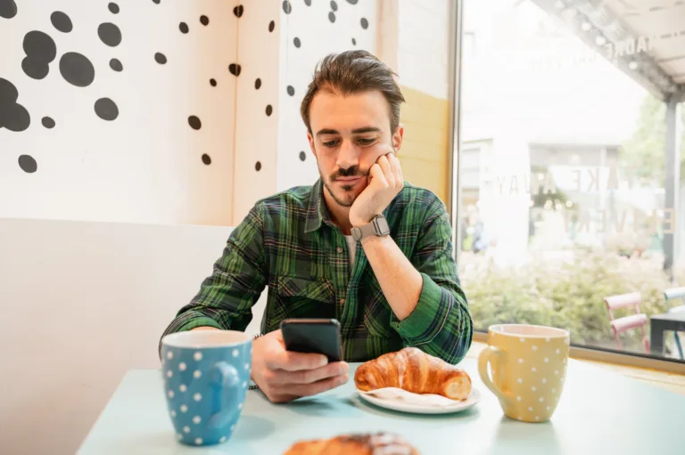 Bearded hipster man sitting at table in a cafe having breakfast. Guy using smartphone.