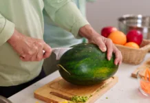 Najpogostejše napake pri rokovanju s hrano, ki lahko ogrozijo vaše zdravje Closeup shot adult hands carefully slicing ripe watermelon with sharp knife on wooden board kitchen counter surrounded by colorful fruits vegetables food prep