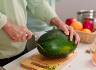 Najpogostejše napake pri rokovanju s hrano, ki lahko ogrozijo vaše zdravje Closeup shot adult hands carefully slicing ripe watermelon with sharp knife on wooden board kitchen counter surrounded by colorful fruits vegetables food prep