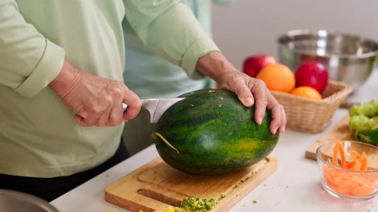 Closeup shot adult hands carefully slicing ripe watermelon with sharp knife on wooden board kitchen counter surrounded by colorful fruits vegetables food prep