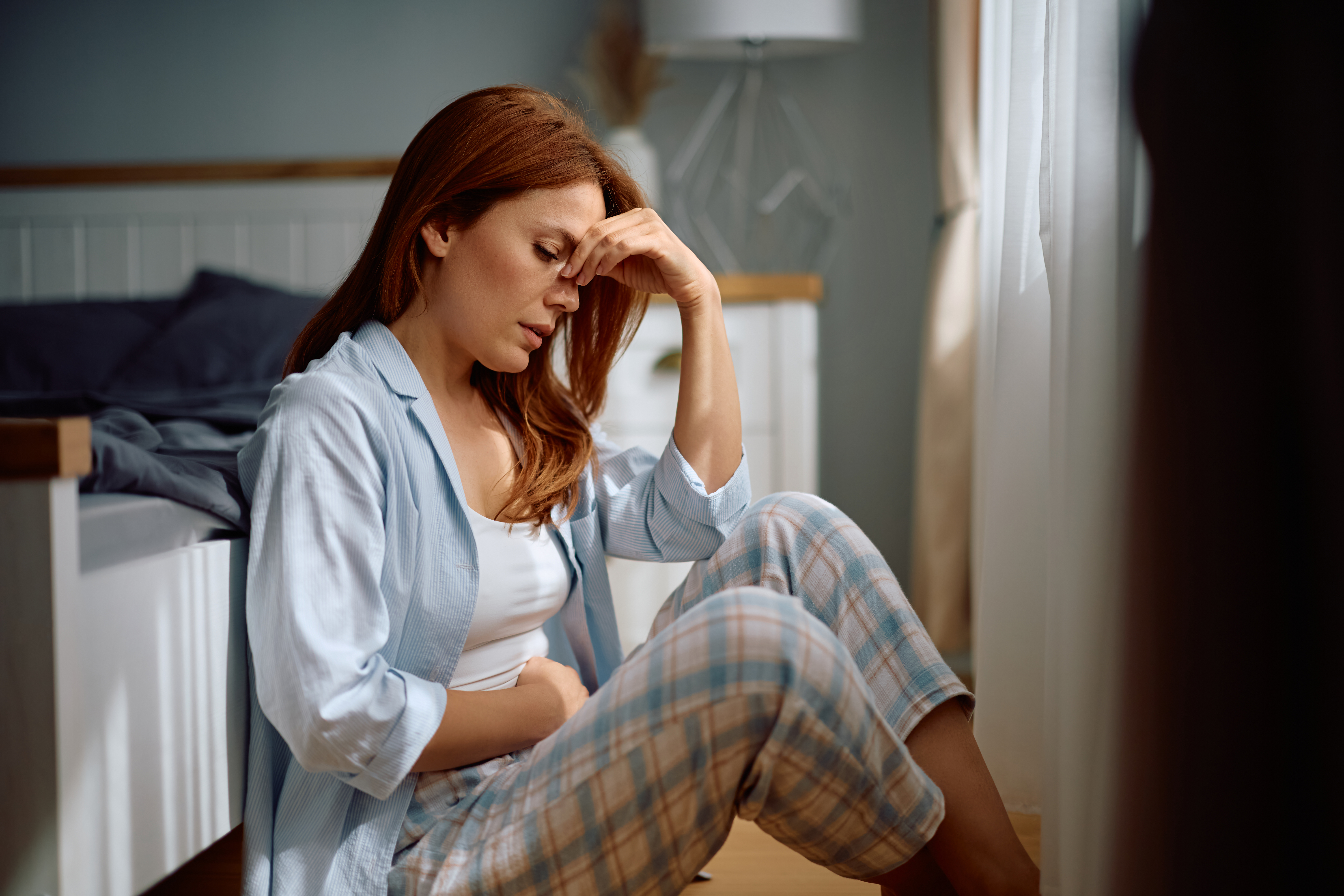 Distraught woman holding her abdomen in pain while sitting on the floor in bedroom.