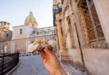 Kje dobro jesti v sicilijanskem Palermu? Hand holding a traditional Sicilian cannolo with ricotta and candied fruit on a historic street in Palermo, Italy. Authentic taste of Mediterranean culture