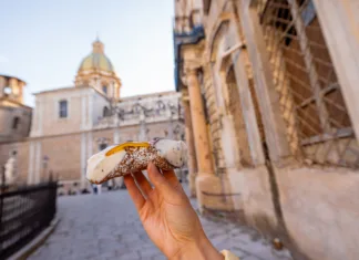Kje dobro jesti v sicilijanskem Palermu? Hand holding a traditional Sicilian cannolo with ricotta and candied fruit on a historic street in Palermo, Italy. Authentic taste of Mediterranean culture