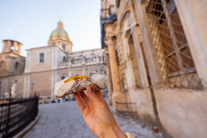 hand-holding-a-traditional-sicilian-cannolo-with-ricotta-and-candied-fruit-on-a-historic-street-in-palermo-italy-authentic-taste-of-mediterranean-culture-stockpack-istock Hand holding a traditional Sicilian cannolo with ricotta and candied fruit on a historic street in Palermo, Italy. Authentic taste of Mediterranean culture