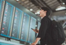 Preživel sem dopust brez dragih agencij in tako svet videl s študentskim proračunom Happy asian man traveller using smartphone checking flight schedule departures board in departure airport terminal hall in front of check in counters. Tourist journey trip concept