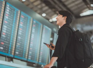 Preživel sem dopust brez dragih agencij in tako svet videl s študentskim proračunom Happy asian man traveller using smartphone checking flight schedule departures board in departure airport terminal hall in front of check in counters. Tourist journey trip concept