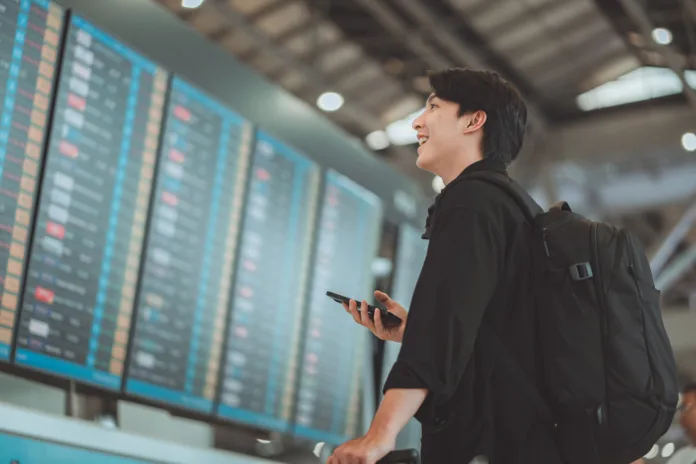 happy-asian-man-traveller-using-smartphone-checking-flight-schedule-departures-board-in-departure-airport-terminal-hall-in-front-of-check-in-counters-tourist-journey-trip-concept-stockpack-istock Happy asian man traveller using smartphone checking flight schedule departures board in departure airport terminal hall in front of check in counters. Tourist journey trip concept