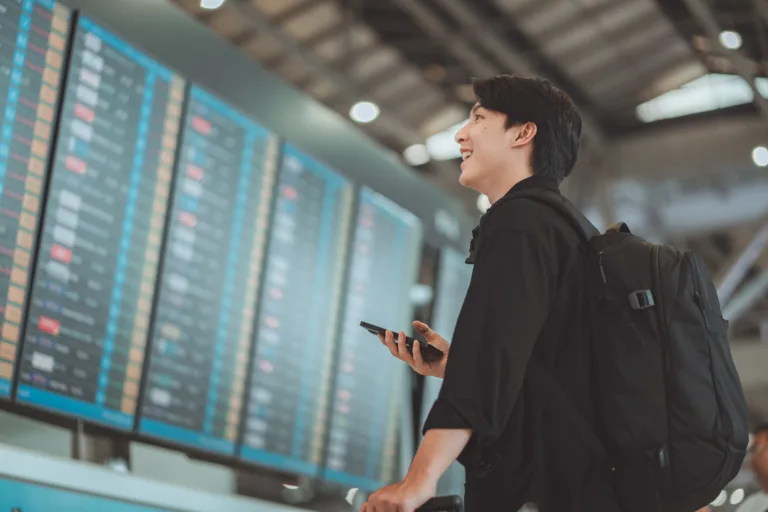 Happy asian man traveller using smartphone checking flight schedule departures board in departure airport terminal hall in front of check in counters. Tourist journey trip concept