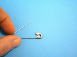Videli ste jo že neštetokrat, a le redki vedo, čemu služi: zato ima vsaka varnostna zaponka to luknjico One metal safety pin positioned on a light blue background with hand and Copy Space, suitable for minimal concepts.