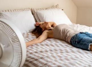 7 skritih trikov, kako ohraniti spalnico hladno Overheated young woman resting on bed using electric fan for relief