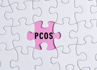 Sindrom policističnih jajčnikov (PCOS) uradno preimenovan v PMOS: Zakaj je to pomembno? PCOS text on white jigsaw puzzle over pink background. Polycystic Ovary Syndrome Health Care concept.