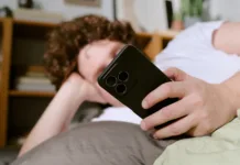 Dopaminski post: Kaj se zgodi, če 48 ur ne gledamo v telefon? Person with curly hair lounging on couch, holding a smartphone, creating a casual and relaxed atmosphere with books and shelves in background