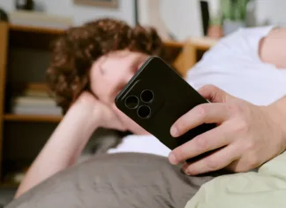 Dopaminski post: Kaj se zgodi, če 48 ur ne gledamo v telefon? Person with curly hair lounging on couch, holding a smartphone, creating a casual and relaxed atmosphere with books and shelves in background