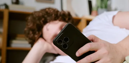 Person with curly hair lounging on couch, holding a smartphone, creating a casual and relaxed atmosphere with books and shelves in background