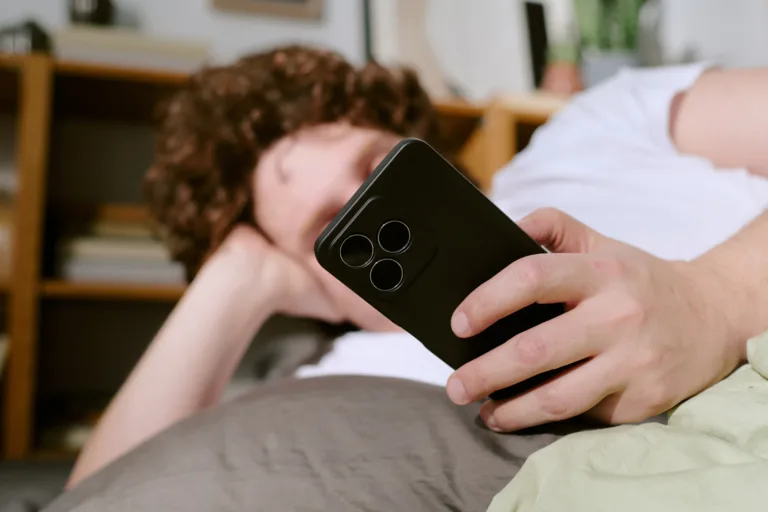 Person with curly hair lounging on couch, holding a smartphone, creating a casual and relaxed atmosphere with books and shelves in background