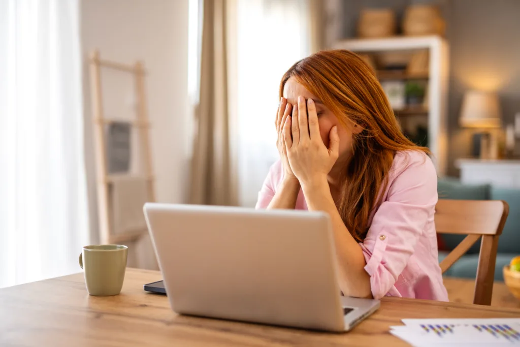Stressed and Tired Young Woman Covering Her Face with Hands While Working on Laptop at Home. Exhausted Female Freelancer Feeling Burnout or Headache Due to Overwork and Stressful Deadline.