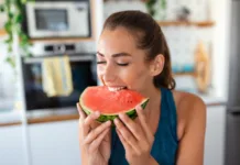 Poleti jo jemo vsi: 5 razlogov, zakaj bi morali lubenico jesti pogosteje Young woman eats a slice of watermelon in the kitchen. Portrait of young woman enjoying a watermelon.
