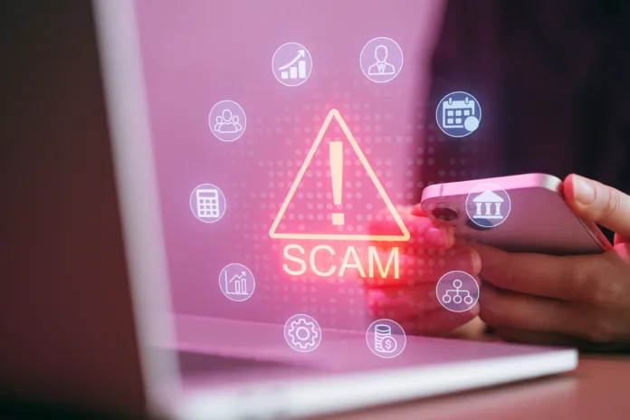 a-person-holds-a-smartphone-with-warning-signs-about-scams-visible-a-laptop-is-nearby-showing-icons-related-to-finance-and-planning-the-setting-is-bright-and-focused-stockpack-istock A person holds a smartphone with warning signs about scams visible. A laptop is nearby, showing icons related to finance and planning. The setting is bright and focused.