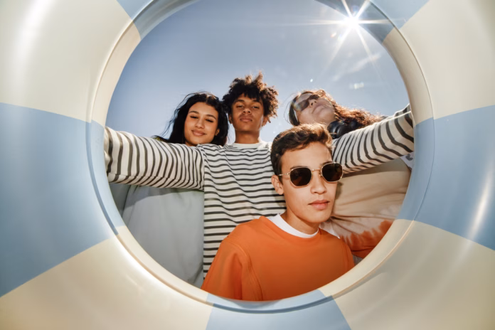 Fisheye shot of four teenagers posing together for camera holding swimming tube while enjoying sunshine on beach, camera flash, copy space