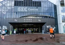 Od 650 do pol milijona tekmovalcev: kako je Hyrox v sedmih letih obrnil fitnes industrijo na glavo SECC, Glasgow, Lanaarkshire, Scotland, United Kingdom - HYROX Glasgow March 2026 event at the SEC Centre - Exterior of the building and entrance, with attendees outside entrance for the fitness racing series. The SEC Centre in Glasgow features a modern glass facade with prominent 'SEC Centre' and 'HYROX GLASGOW' signage. The building displays 'THE WORLD SERIES OF FITNESS RACING' above its main entrance. People gather outside the venue. Staff in high-visibility vests assist at the entrance. The ground consists of wet, reddish-brown paving stones with puddles, indicating recent rain. Branding features 'HYROX' text and fitness-themed murals on the facade.