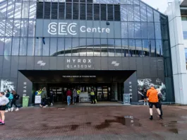 Od 650 do pol milijona tekmovalcev: kako je Hyrox v sedmih letih obrnil fitnes industrijo na glavo SECC, Glasgow, Lanaarkshire, Scotland, United Kingdom - HYROX Glasgow March 2026 event at the SEC Centre - Exterior of the building and entrance, with attendees outside entrance for the fitness racing series. The SEC Centre in Glasgow features a modern glass facade with prominent 'SEC Centre' and 'HYROX GLASGOW' signage. The building displays 'THE WORLD SERIES OF FITNESS RACING' above its main entrance. People gather outside the venue. Staff in high-visibility vests assist at the entrance. The ground consists of wet, reddish-brown paving stones with puddles, indicating recent rain. Branding features 'HYROX' text and fitness-themed murals on the facade.