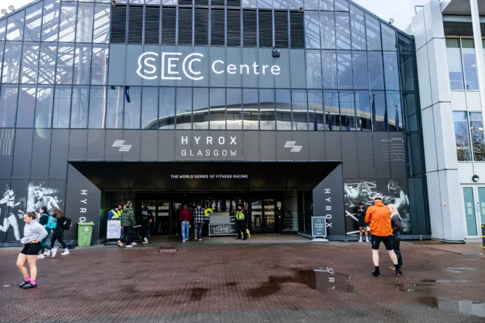 secc-glasgow-lanaarkshire-scotland-united-kingdom-hyrox-glasgow-march-2026-event-at-the-sec-centre-exterior-of-the-building-and-entrance-with-attendees-outside-entrance-for-the-fitness-racing-series-t-stockpack-istock SECC, Glasgow, Lanaarkshire, Scotland, United Kingdom - HYROX Glasgow March 2026 event at the SEC Centre - Exterior of the building and entrance, with attendees outside entrance for the fitness racing series. The SEC Centre in Glasgow features a modern glass facade with prominent 'SEC Centre' and 'HYROX GLASGOW' signage. The building displays 'THE WORLD SERIES OF FITNESS RACING' above its main entrance. People gather outside the venue. Staff in high-visibility vests assist at the entrance. The ground consists of wet, reddish-brown paving stones with puddles, indicating recent rain. Branding features 'HYROX' text and fitness-themed murals on the facade.