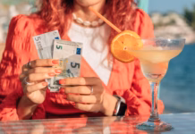 Dopust postaja luksuz? Skoraj vsak tretji Evropejec si tedna počitnic ne more privoščiti Woman sitting at a beach cafe, holding Euro banknotes and counting cash with a smile, cocktail on the table, vacation setting